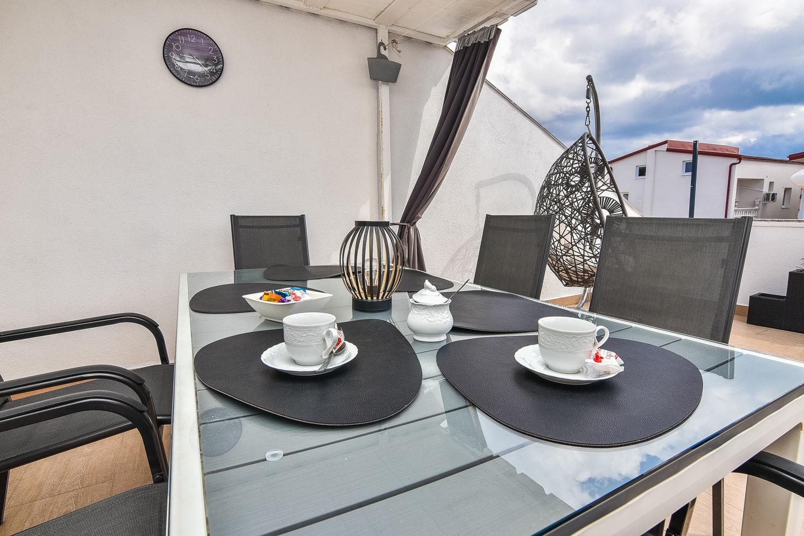Terrasse mit Glastisch, Stühlen und Hängesessel. Esstisch mit Tassen und Untertassen.