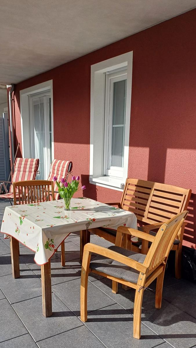 Terrasse mit Holztisch, Stühlen und Blumen. Rote Wand und Fenster.
