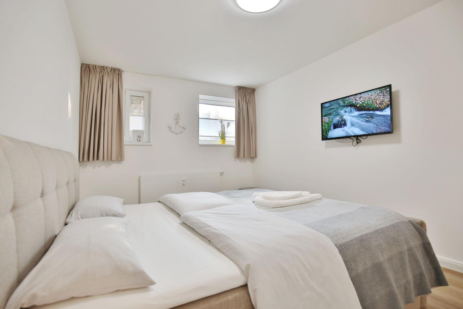 Schlafzimmer mit Doppelbett, TV und zwei Fenstern mit Vorhängen.