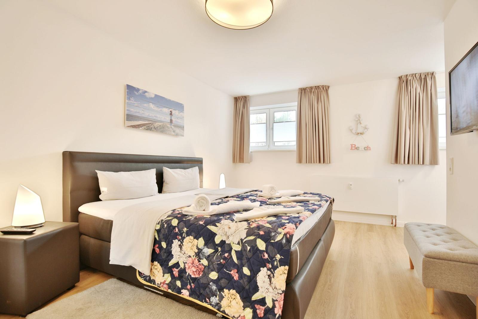 Schlafzimmer mit Bett, Nachttisch, Fernseher und Fenster mit Vorhängen.