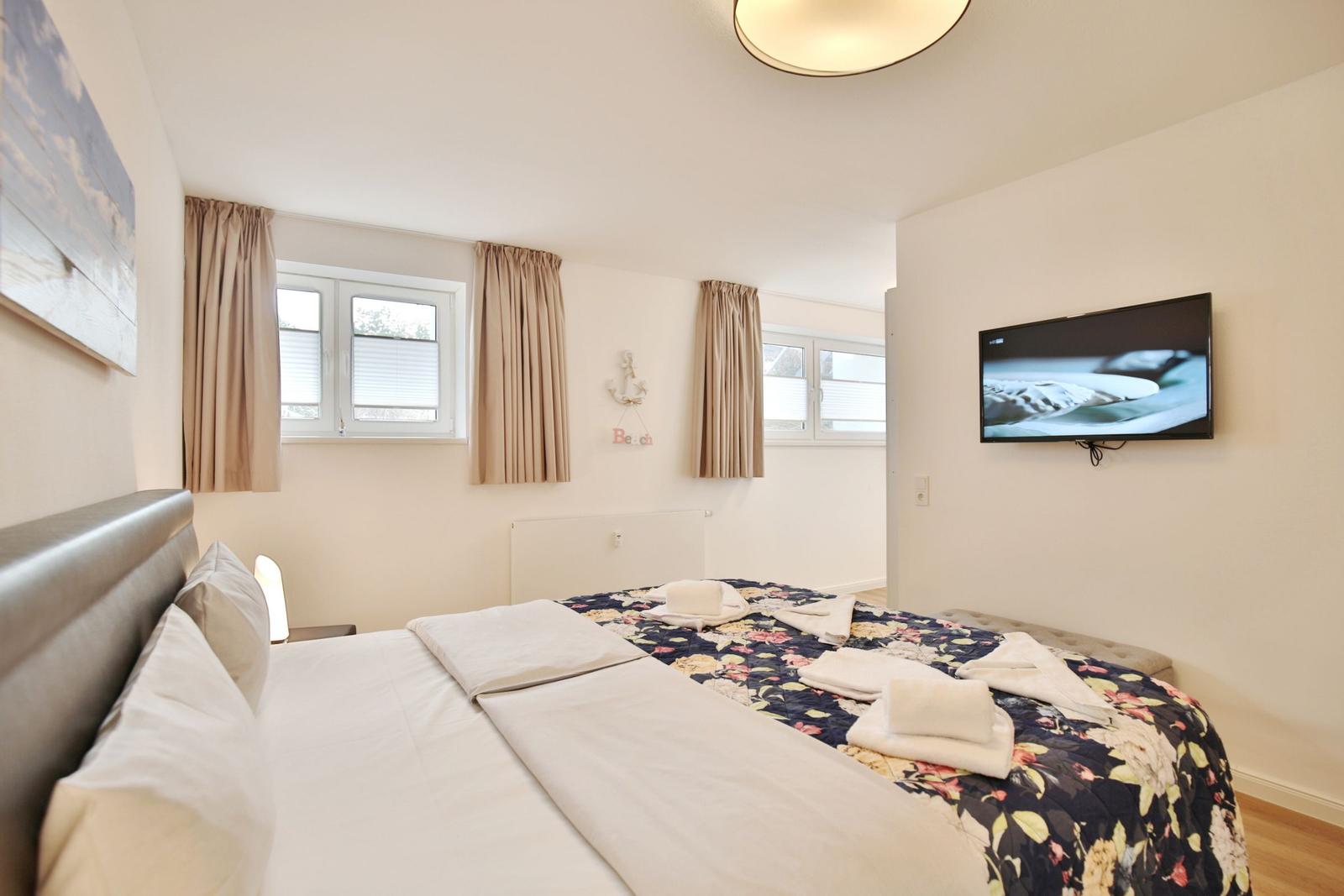 Schlafzimmer mit Bett, TV und Fenstern mit Vorhängen.