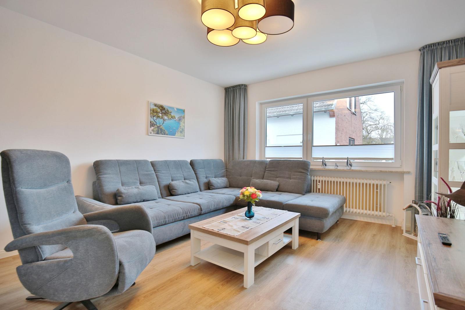 Wohnzimmer mit grauem Sofa, Sessel und Holztisch. Fenster mit Jalousien und Wandbild.