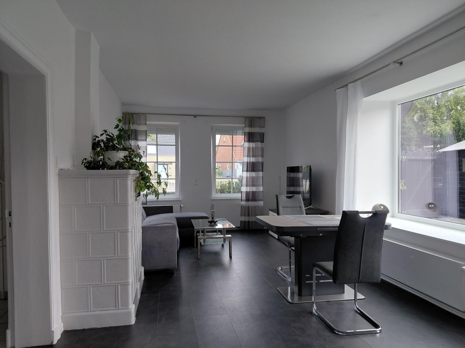 Wohn- und Essbereich mit Sofa, Tisch und Stühlen. Große Fenster mit Blick nach draußen.