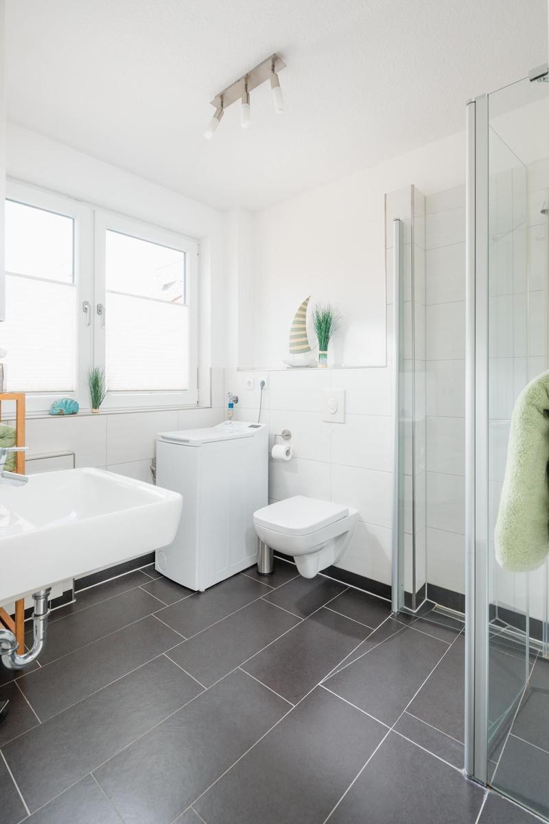 Badezimmer mit Dusche, Waschbecken und Toilette. Fenster, graue Fliesen und weiße Wände.