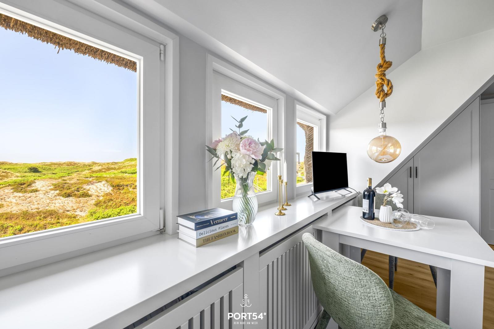 Schreibtisch mit Sicht auf Dünenlandschaft, Blumen, Bücher und Lampe.