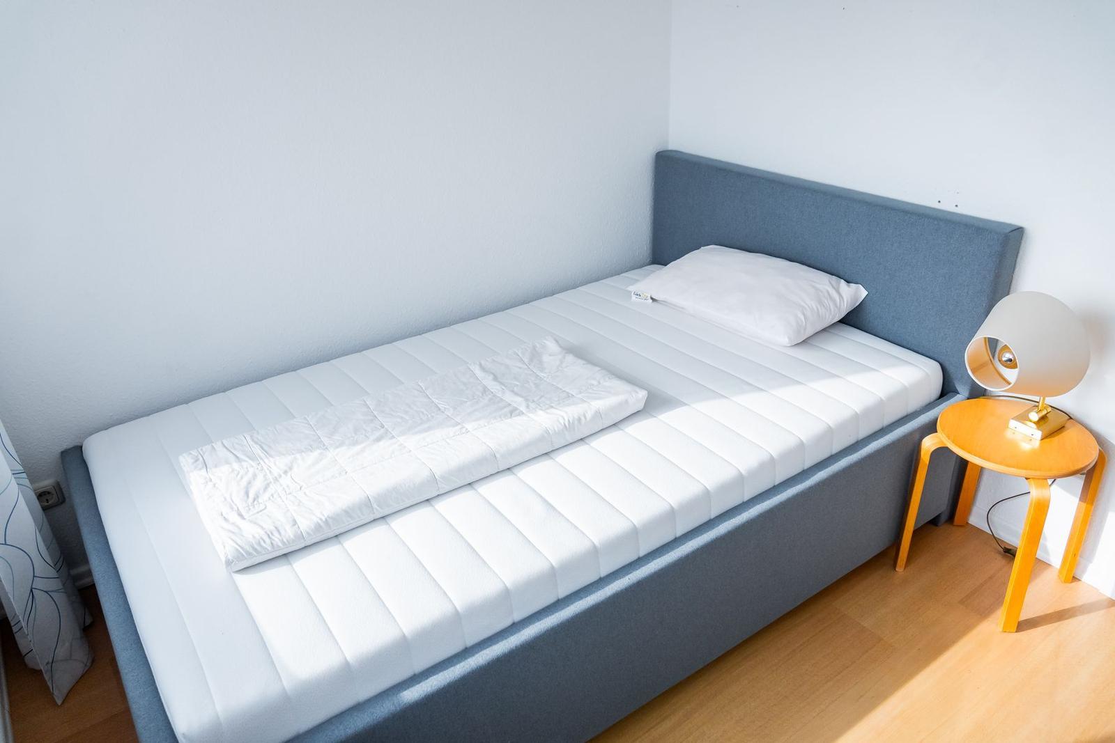 Ein Bett mit weißer Matratze und Kissen. Neben dem Bett steht ein kleiner Tisch mit einer Lampe.