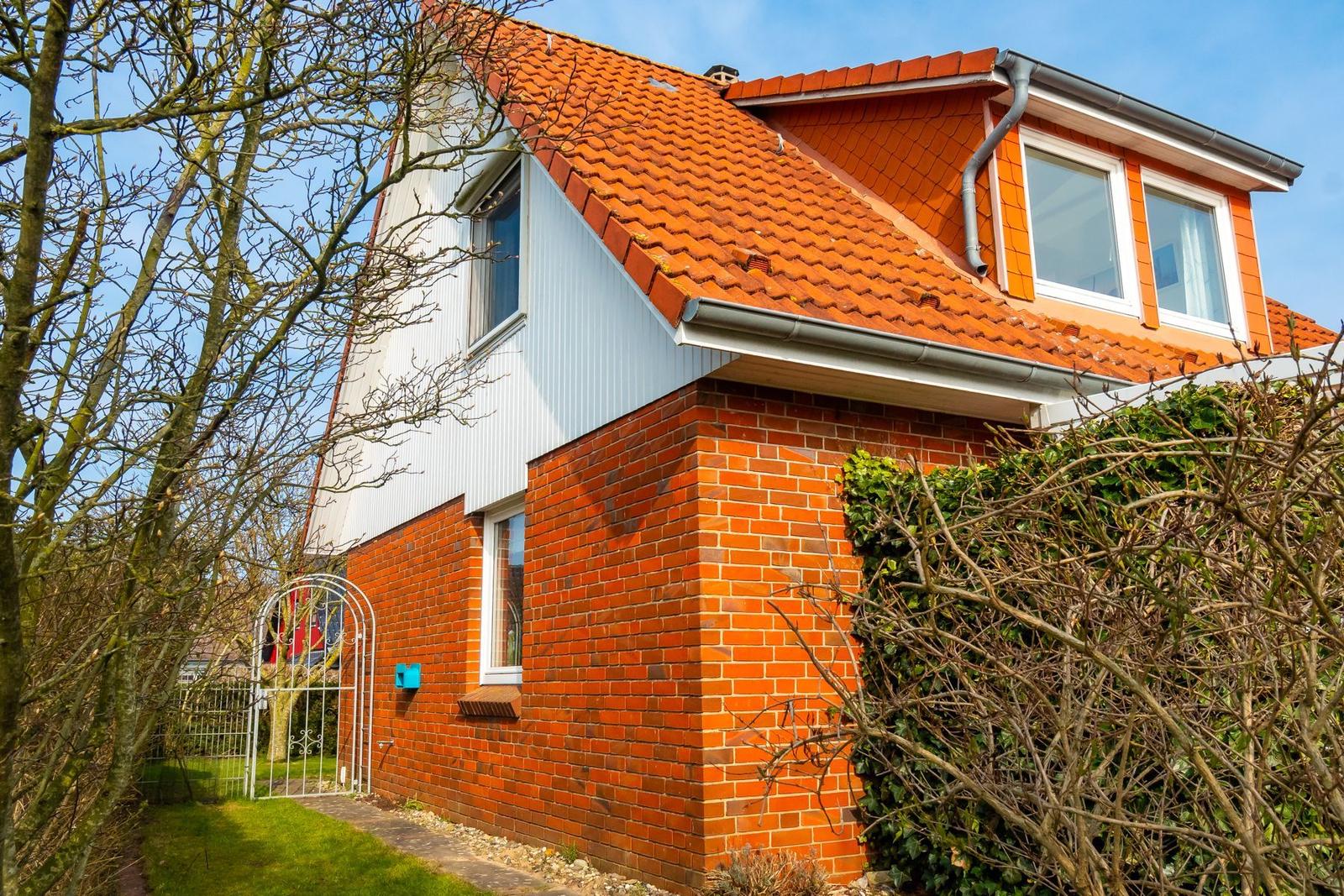 Haus mit rotem Ziegeldach, Backsteinmauer und weißen Fenstern. Eingangstor und Beet sind sichtbar.