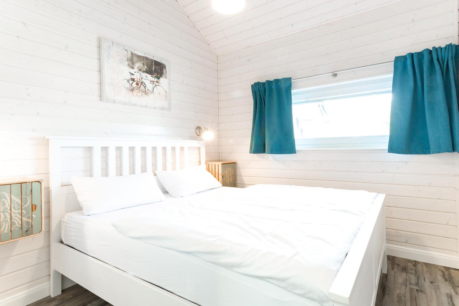 Schlafzimmer mit weißem Bett, Holzwänden und blauen Vorhängen.