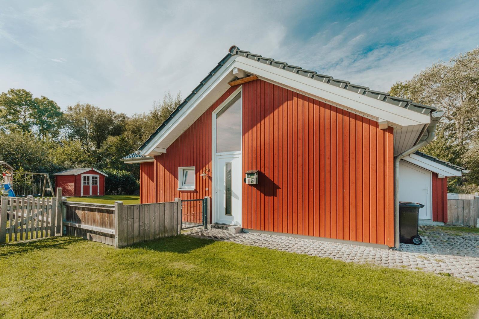 Rotes Holzhaus mit weißer Tür und Garten. Spielplatz und kleiner Schuppen im Hintergrund.