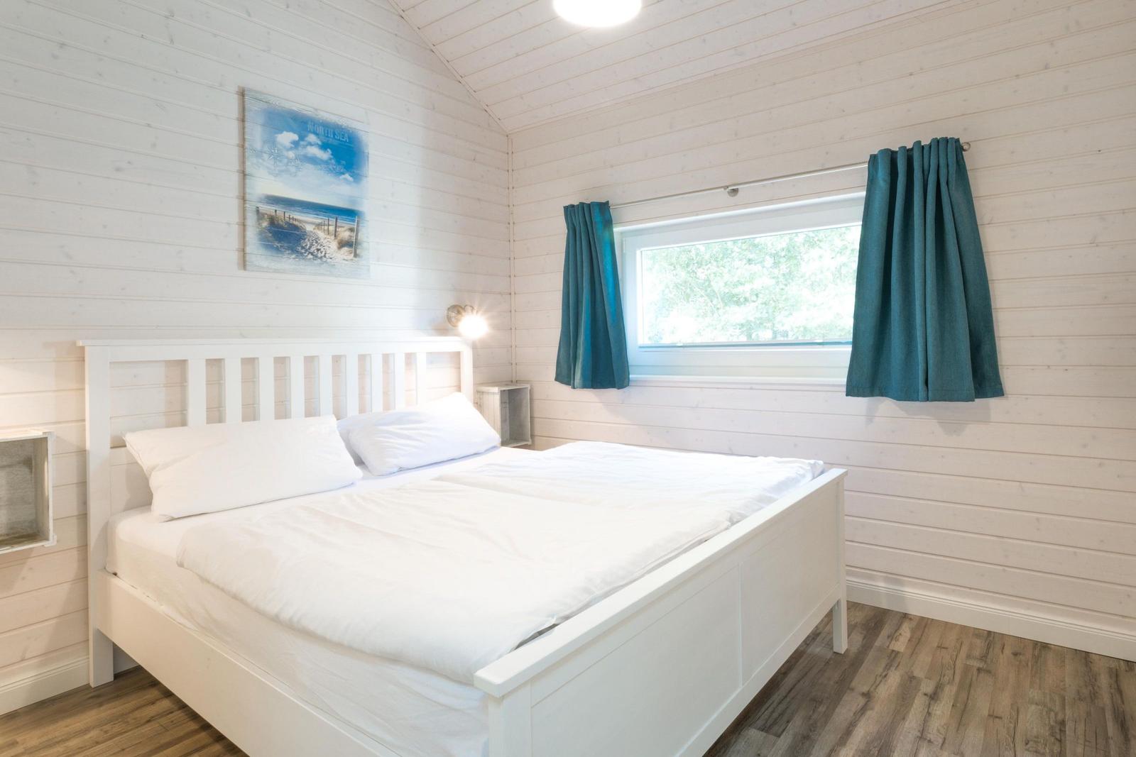 Schlafzimmer mit weißem Bett, Holzwand und Fenster mit blauen Vorhängen.