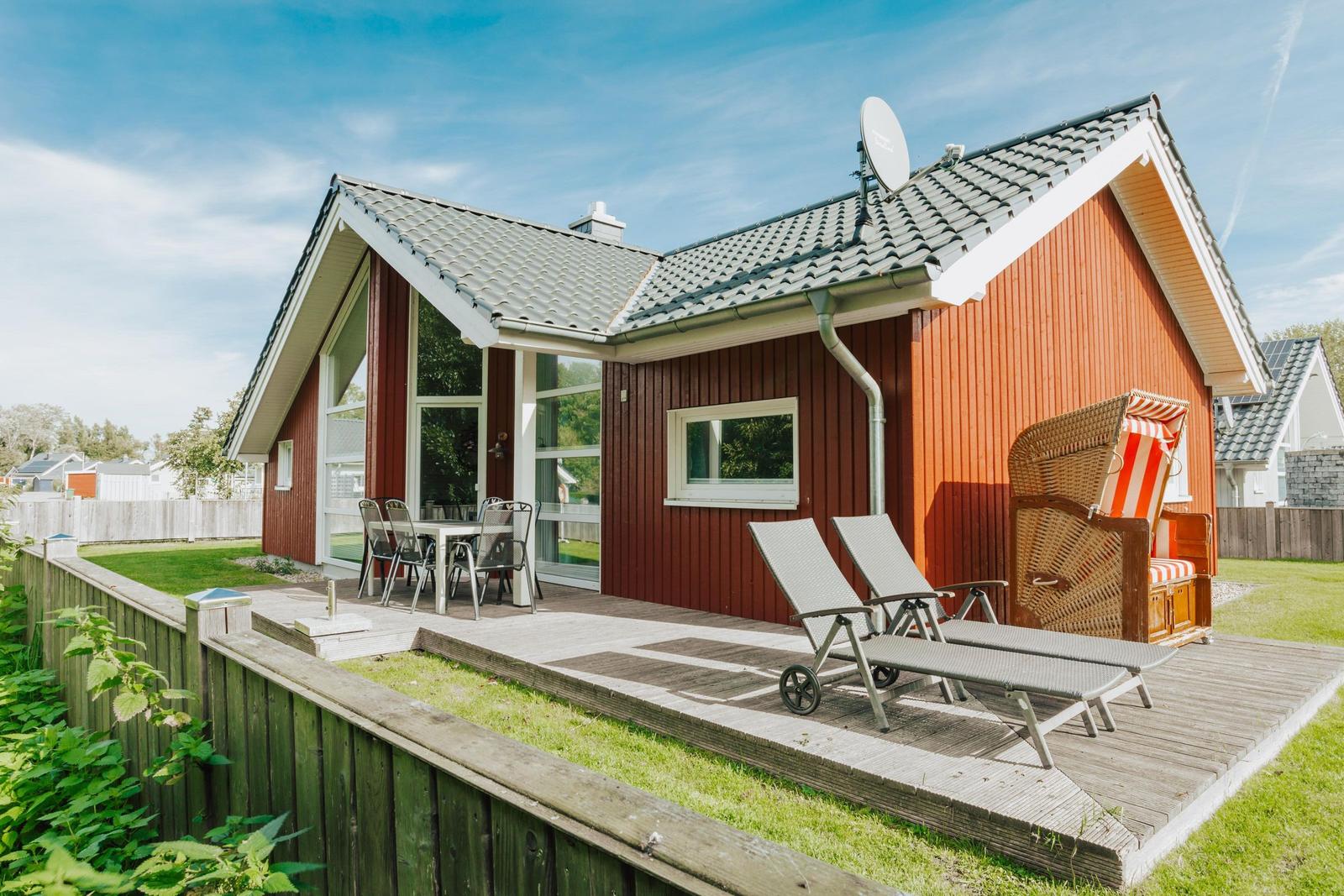 Rotes Ferienhaus mit Terrasse, Liegen und Strandkorb. Grün umgeben.