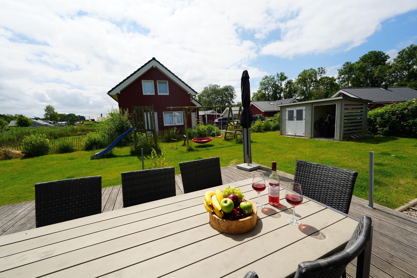 Holztisch mit Obst und Wein auf Terrasse vor rotem Haus mit Spielplatz.