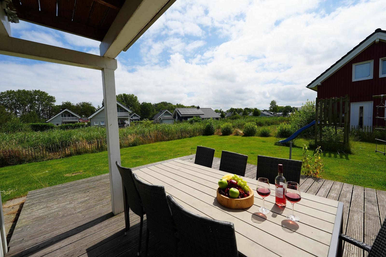 Terrasse mit Tisch, Stühlen, Obst und Wein. Blick auf grünen Garten und roten Haus.