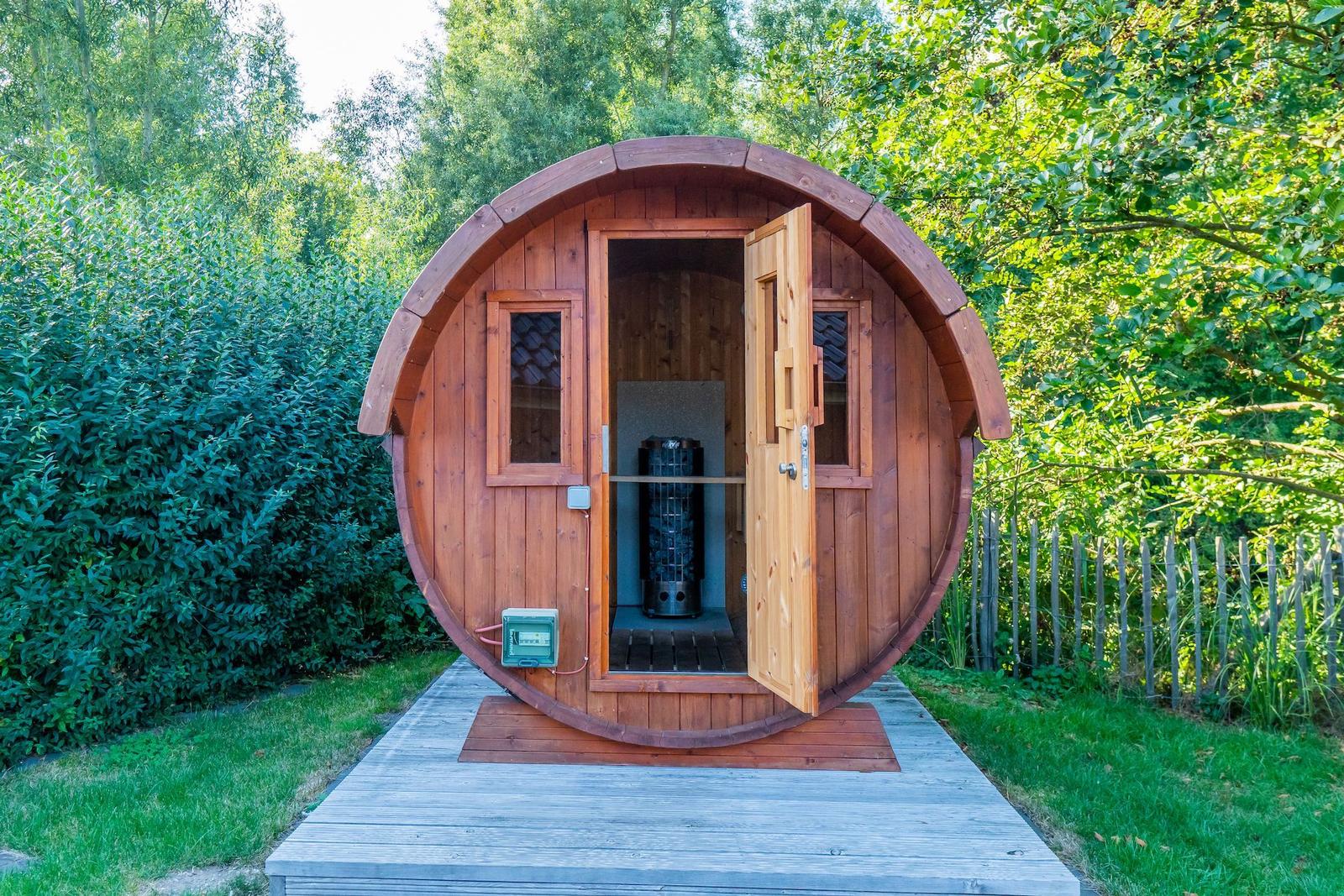 Holzsauna im Stil eines Fasses auf einer Holzterrasse umgeben von Grün.
