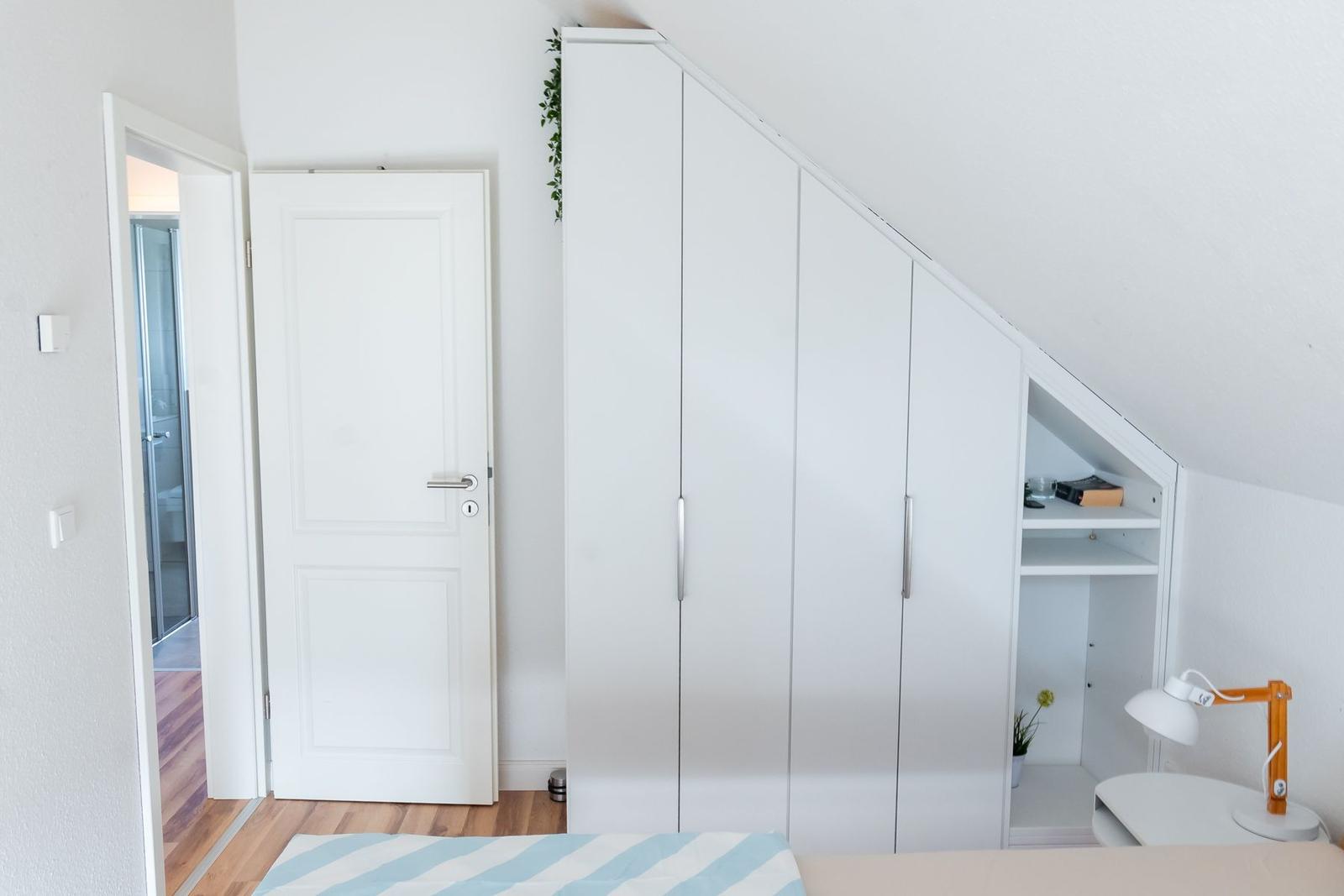 Schlafzimmer mit weißem Schrank und Bett unter Dachschräge. Tür führt in Bad.