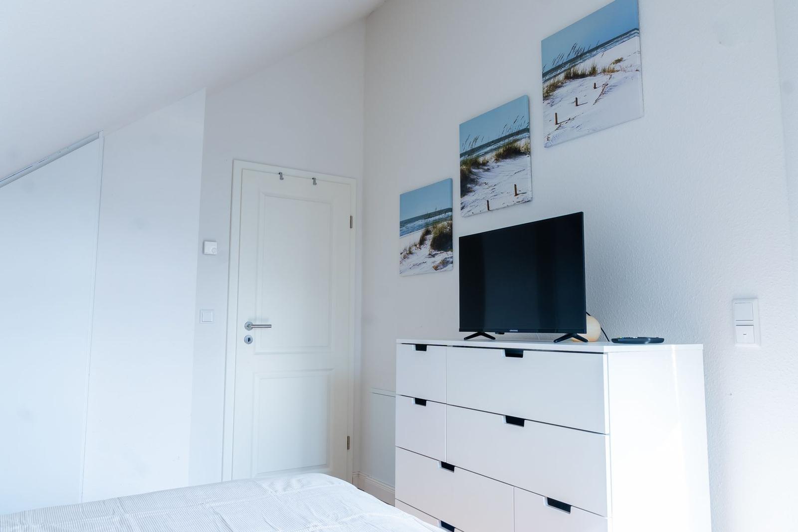 Schlafzimmer mit weißem Schrank, Fernseher und Strandbildern an der Wand.