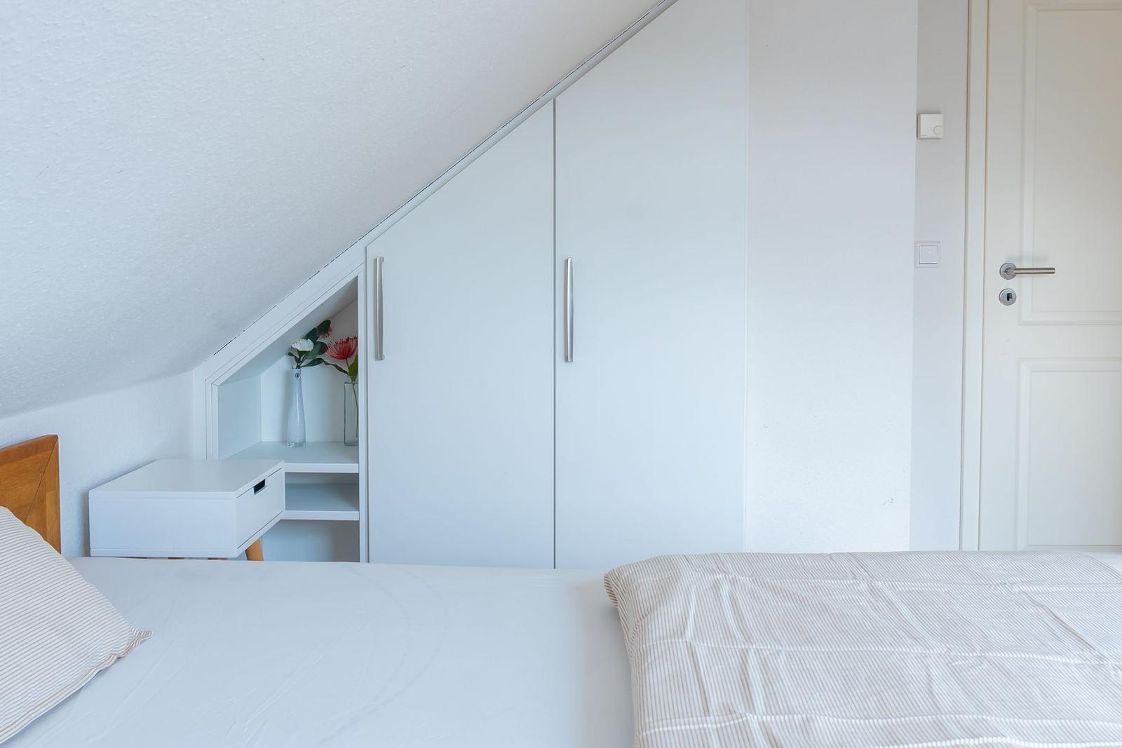 Schlafzimmer mit Dachschräge, weißem Schrank und Nachttisch mit Blumen.