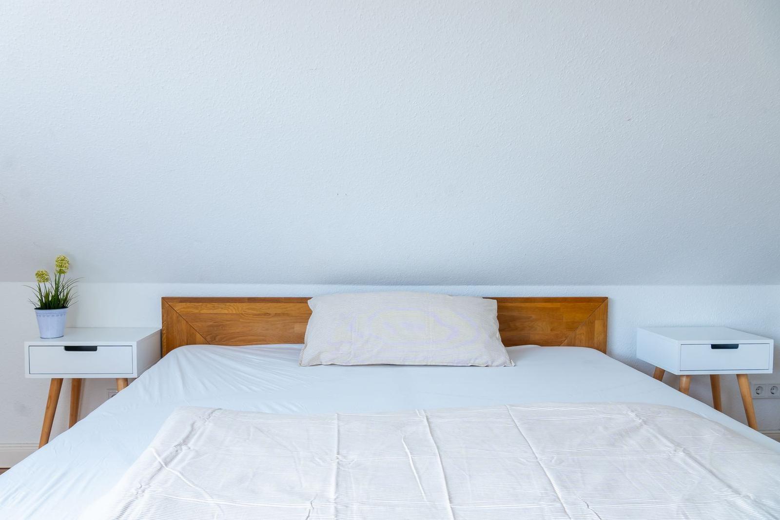 Schlafzimmer mit Bett, Holz-Kopfteil und zwei weißen Nachttischen mit Pflanze.