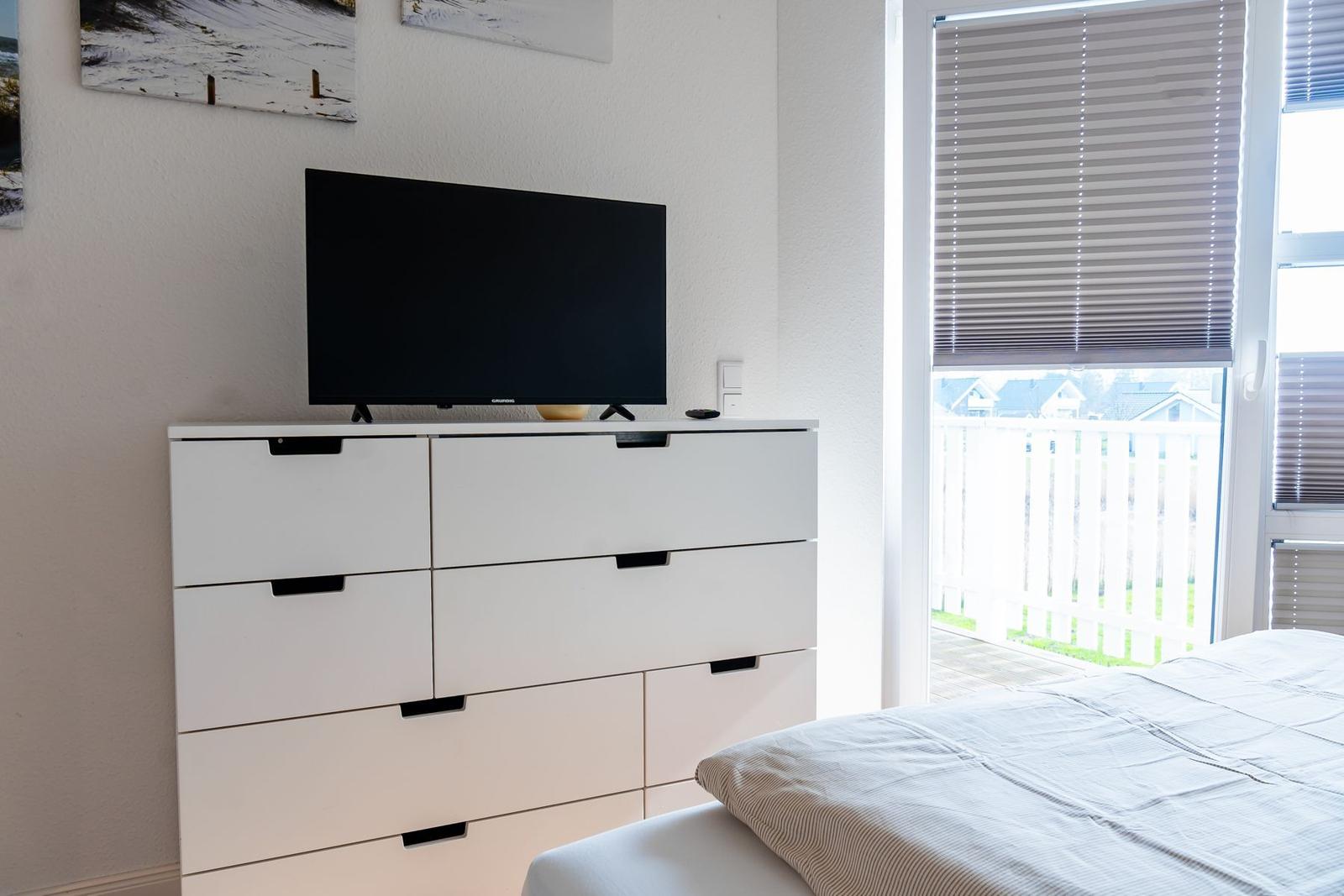 Schlafzimmer mit Bett, Schrank und Fernseher. Fenster mit Jalousie und Blick auf Balkon.