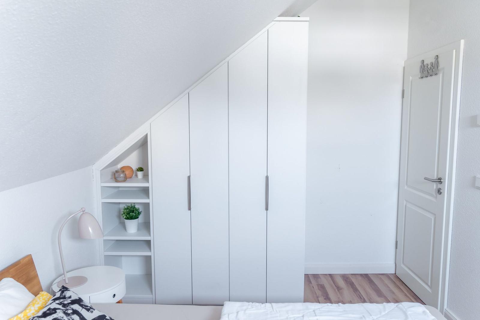 Schlafzimmer mit weißem Schrank und Holzboden. An der Wand befindet sich ein kleiner Regalschrank.