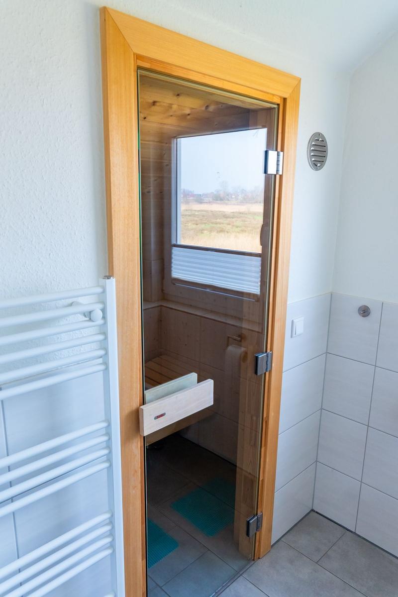 Sauna mit Holzwand und Fenster, Tür mit Glas, daneben Heizung und Fliesenwand.