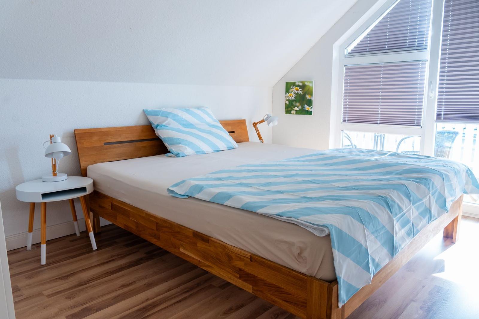 Schlafzimmer mit Holzbett, blau-weißem Bettzeug und großem Fenster mit Jalousien.