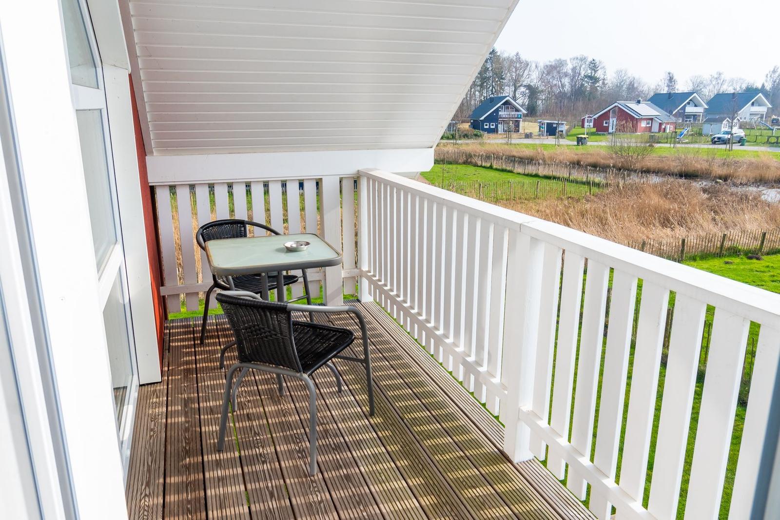 Terrasse mit Tisch und zwei Stühlen. Weißes Geländer und Holzboden. Blick auf Grünland und Häuser.