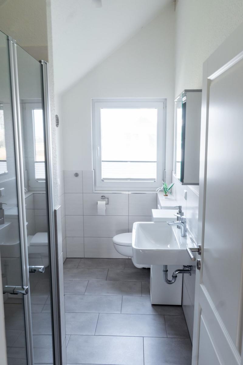 Badezimmer mit Dusche, Waschbecken und Toilette. Fenster sorgt für Tageslicht.