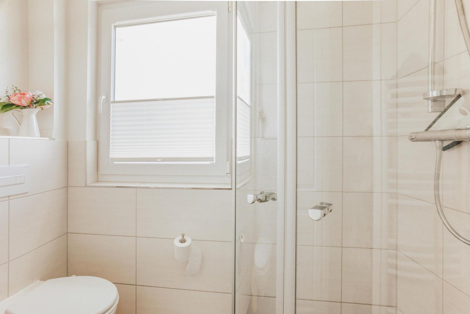 Badezimmer mit Dusche, Toilette und Fenster mit Jalousie.