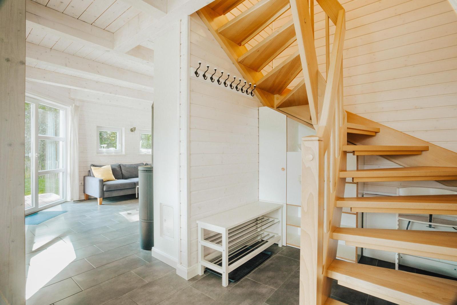 Eingangsbereich mit Holztreppe und Garderobe. Im Hintergrund Wohnzimmer mit Sofa und Fenster.