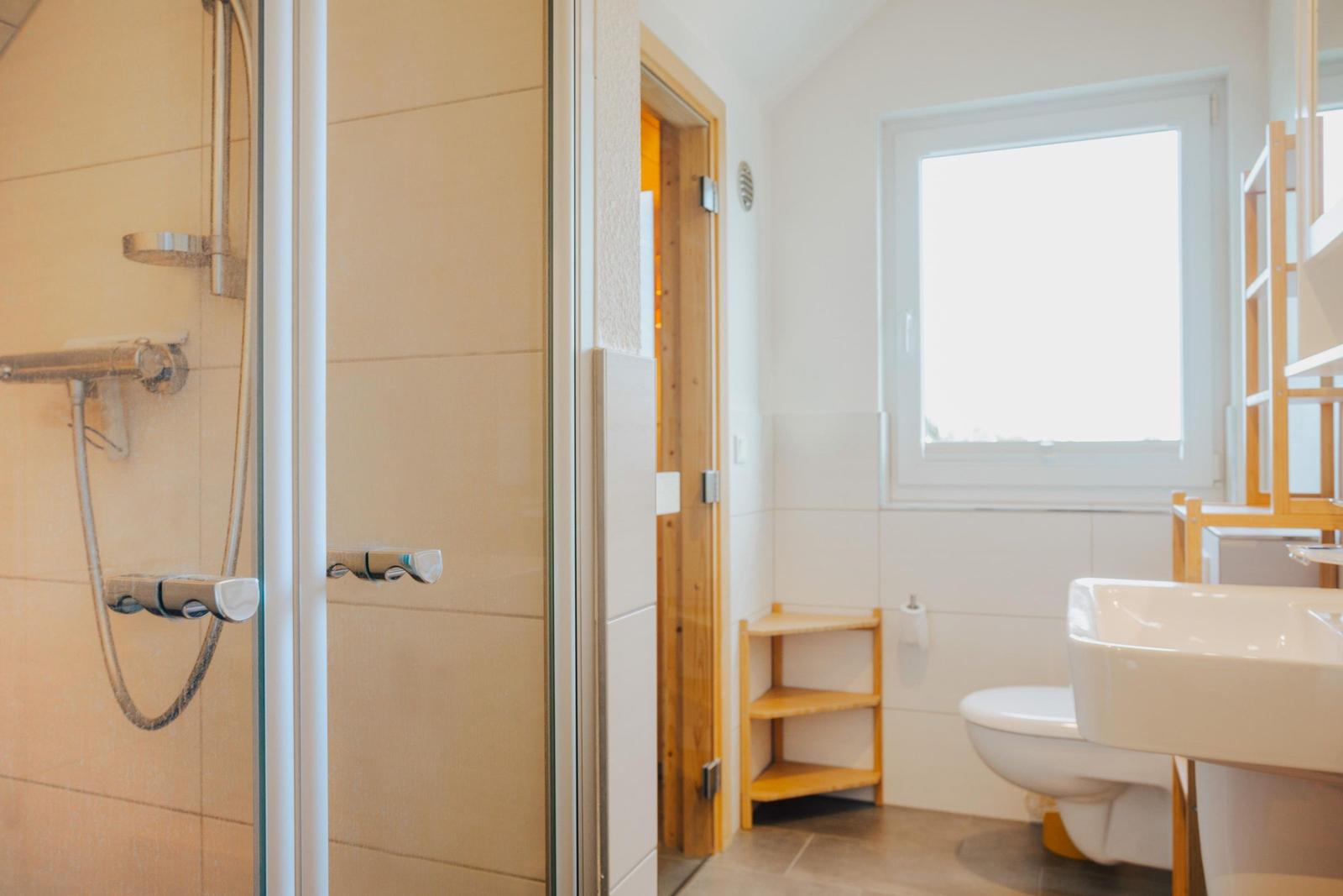 Bad mit Dusche, Waschbecken und Toilette. Holzregal und Fenster sichtbar.