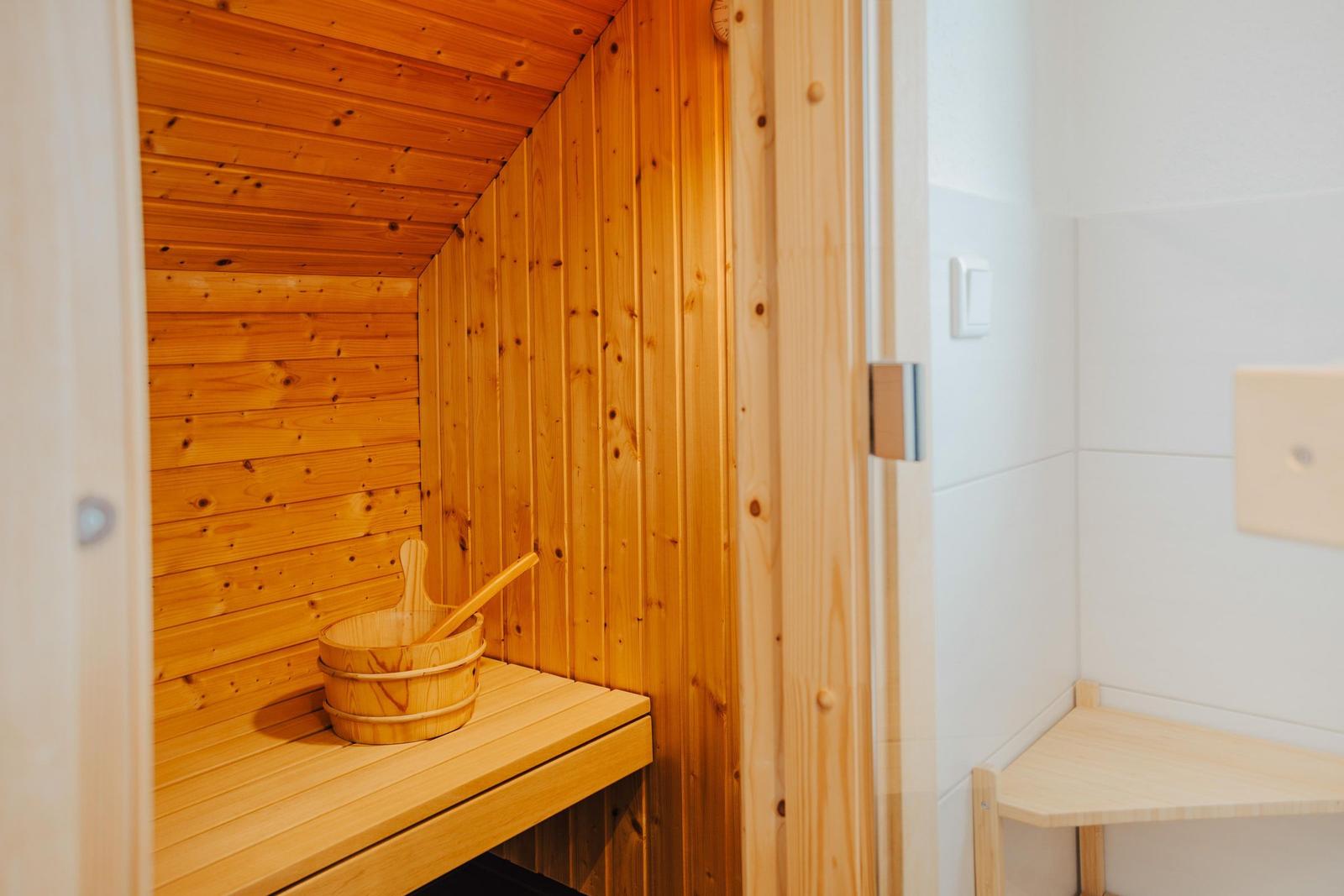 Holzsauna mit Sitzbank und Eimer. Türöffnung zeigt weiße Wand.