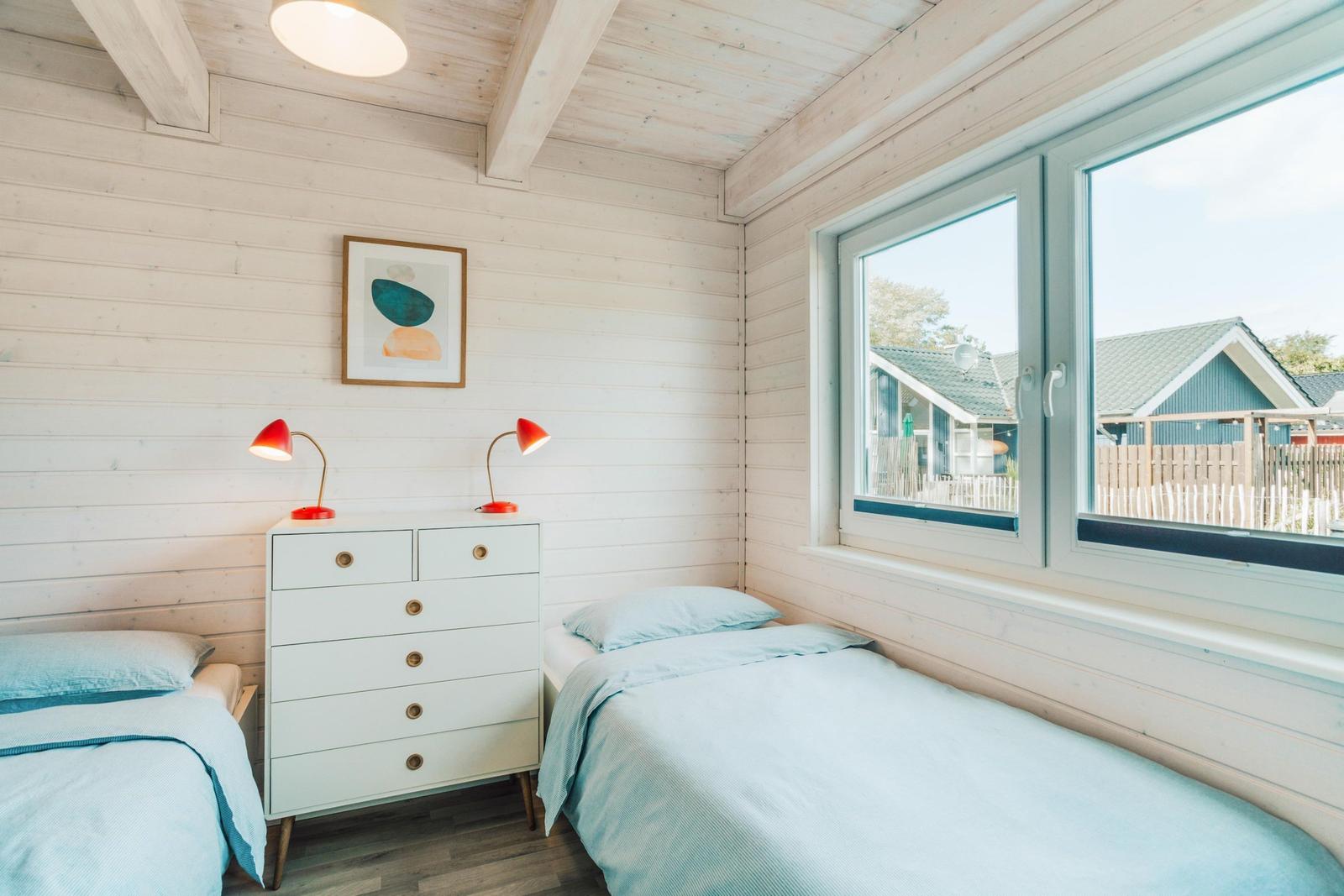 Zwei Betten mit blauen Bettwäsche, weißer Kommode und roten Lampen. Holzwände und große Fenster.