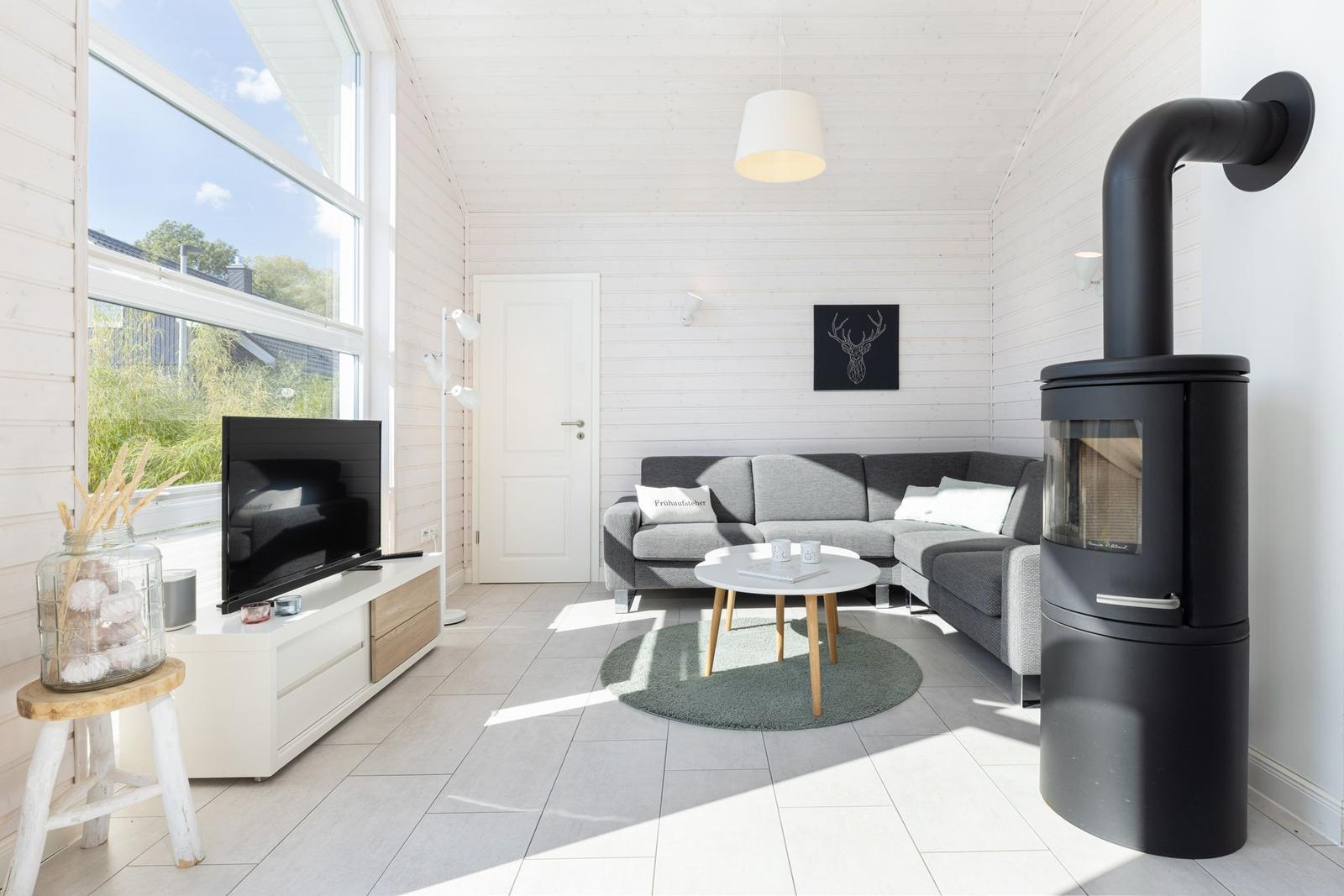 Wohnzimmer mit grauem Sofa, Holztisch und Kaminofen. Große Fenster und weißer Holzboden.