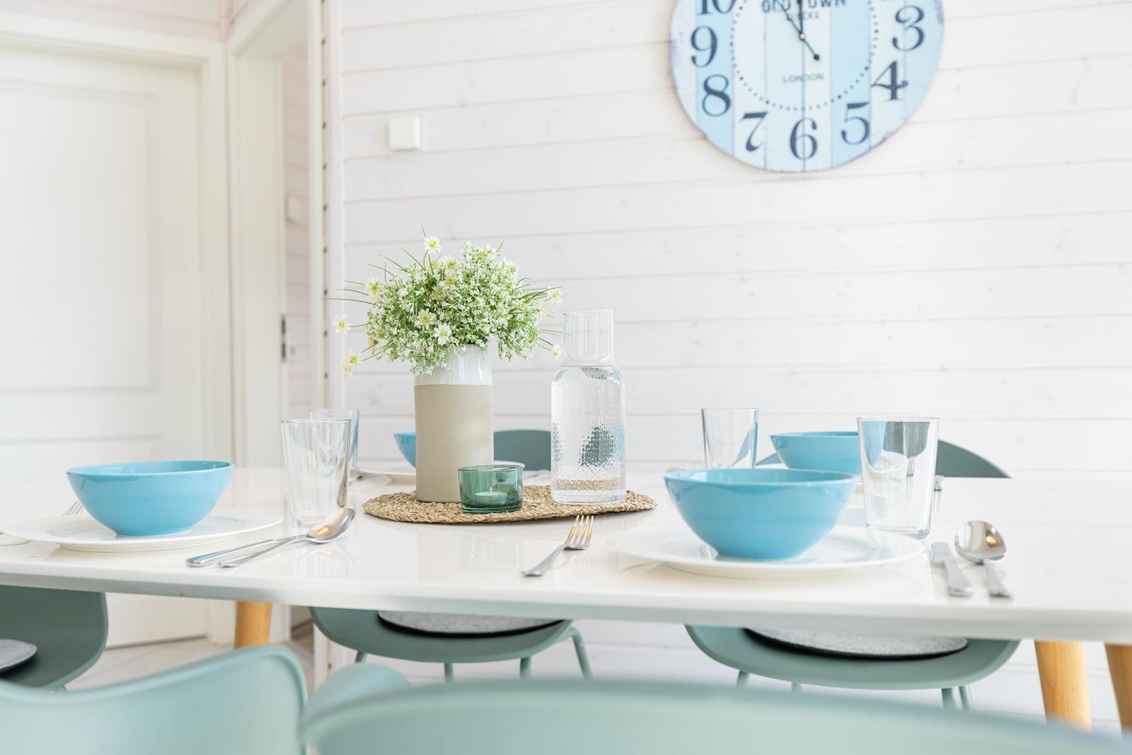 Esstisch mit blauen Schalen und Vasenblumen an weißer Wand mit Wanduhr.