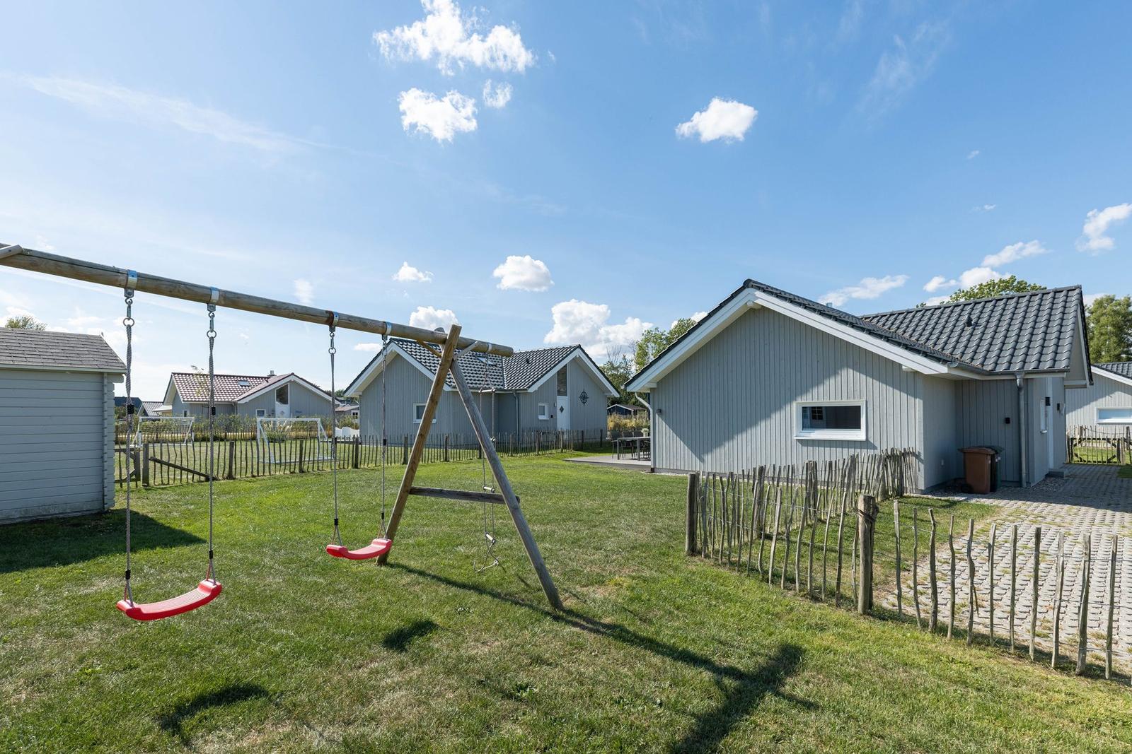 Rotes Schaukeln im Garten vor einem Ferienhaus mit Holzzaun und grünem Rasen.