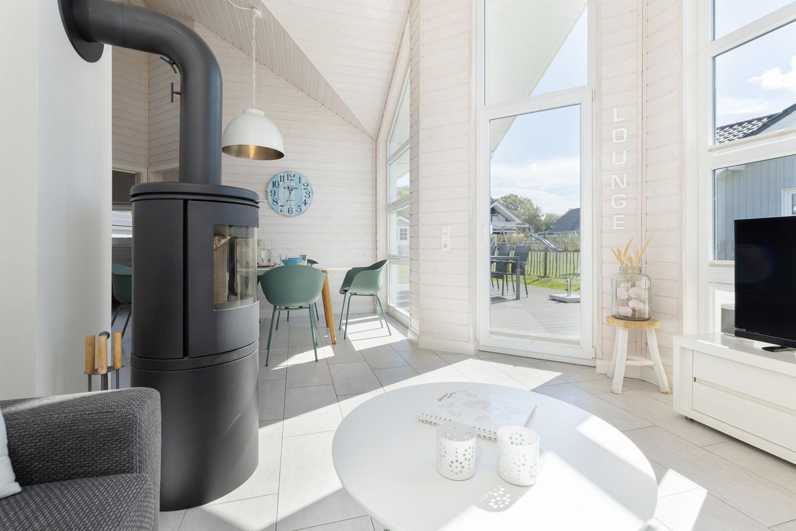 Lounge mit Holzwand, Glasfront und Kaminofen. Tisch, Stühle und TV sichtbar.