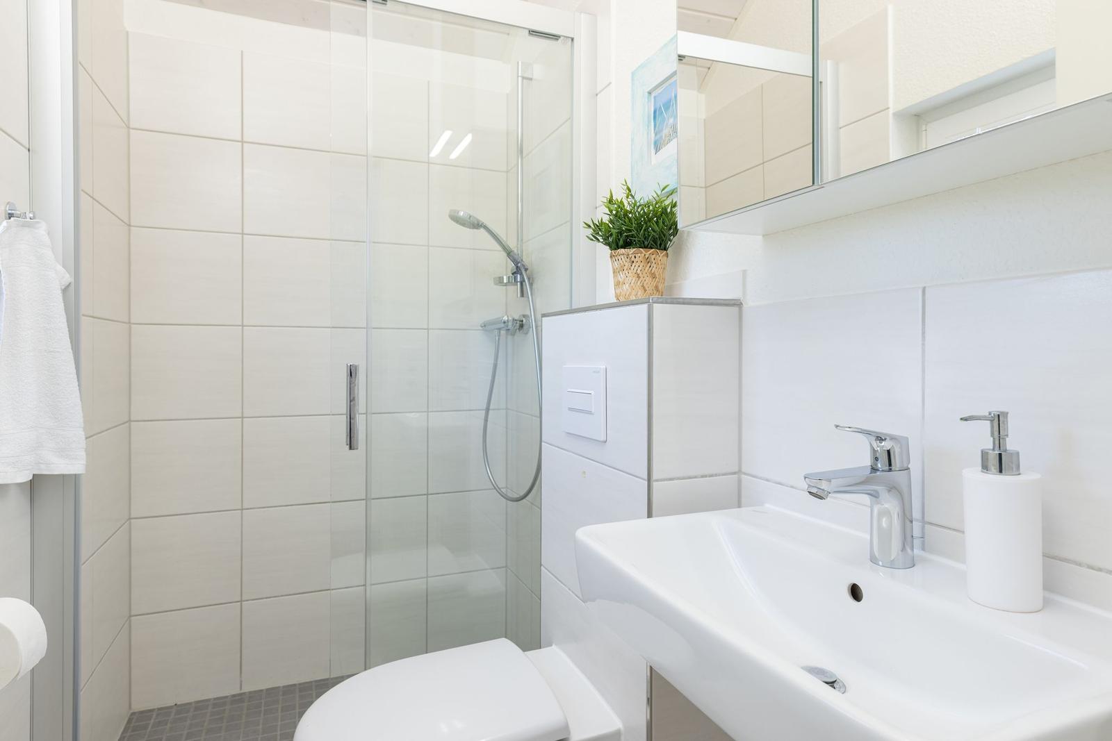 Badezimmer mit Dusche, Waschbecken und Toilette. Weiße Fliesen und Spiegelschrank.