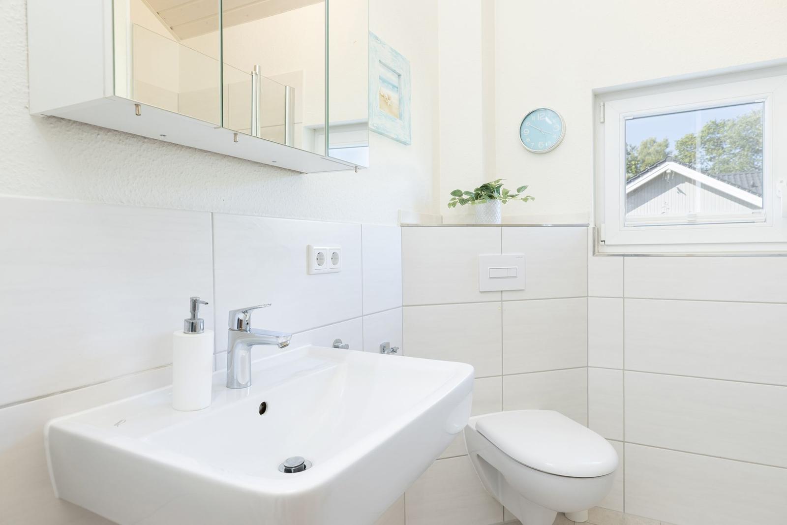 Badezimmer mit Waschbecken, Toilette und Fenster. Spiegel, Pflanze und Uhr sind sichtbar.