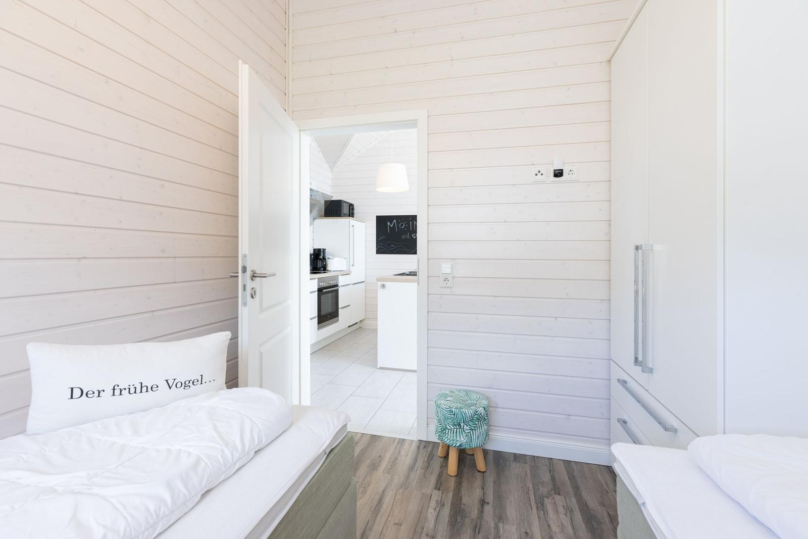 Zimmer mit Bett, Tür zum Küchenbereich und Stuhl. Holzwände und weißer Schrank.