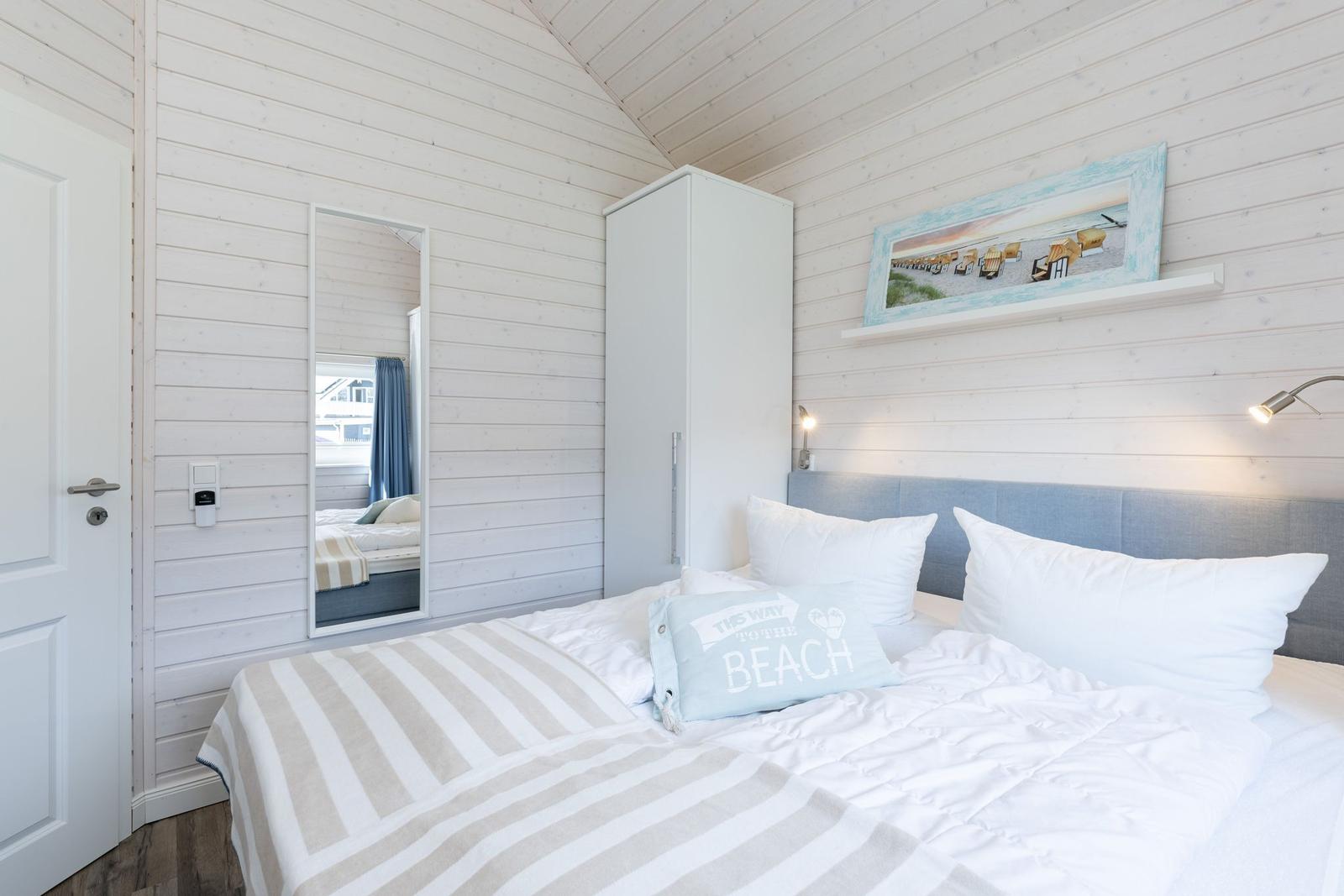 Schlafzimmer mit Bett, Kleiderschrank und Spiegel an weißer Holzwand.