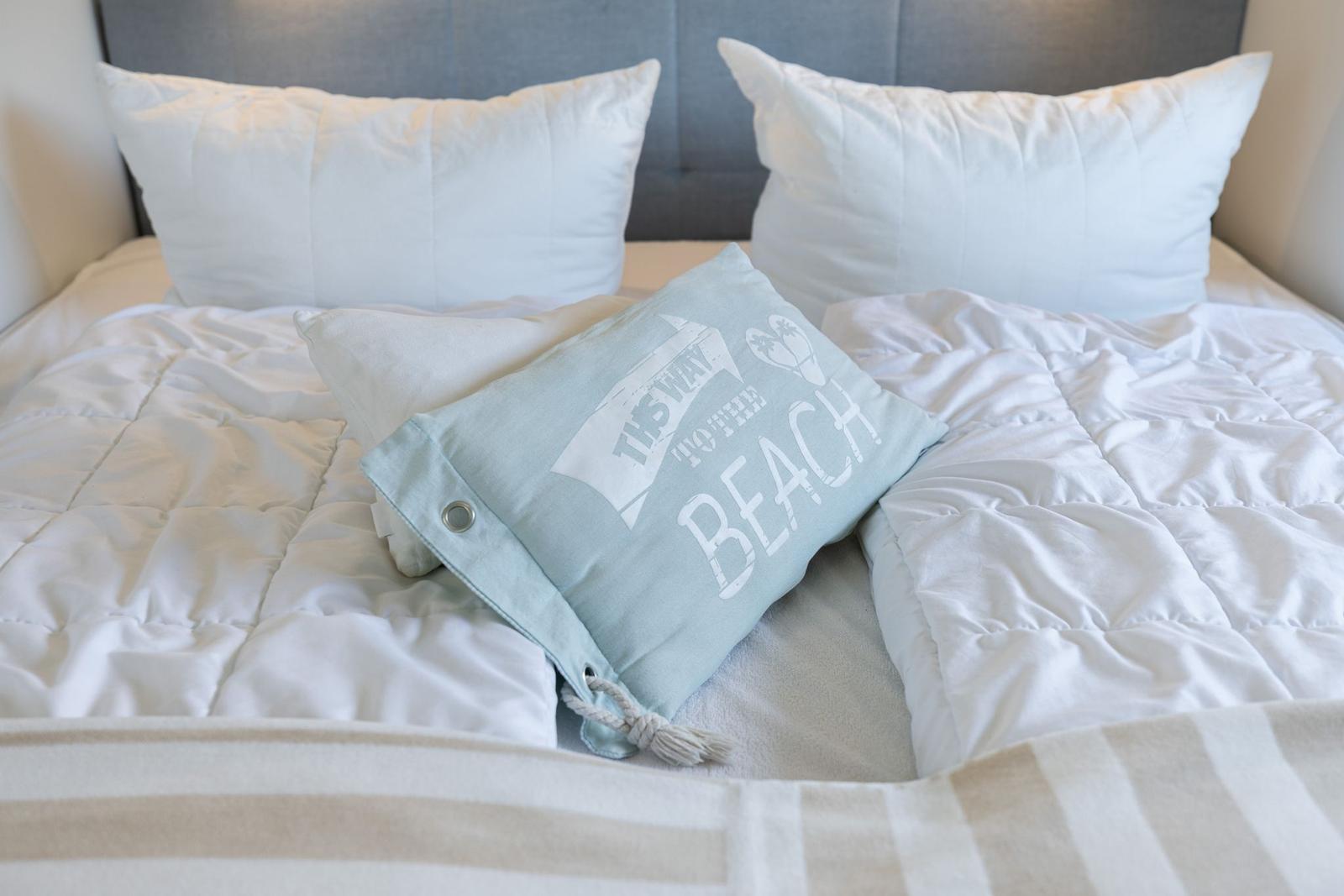 Doppelbett mit weißer Bettwäsche und einem hellblauen Kissen mit Aufschrift.