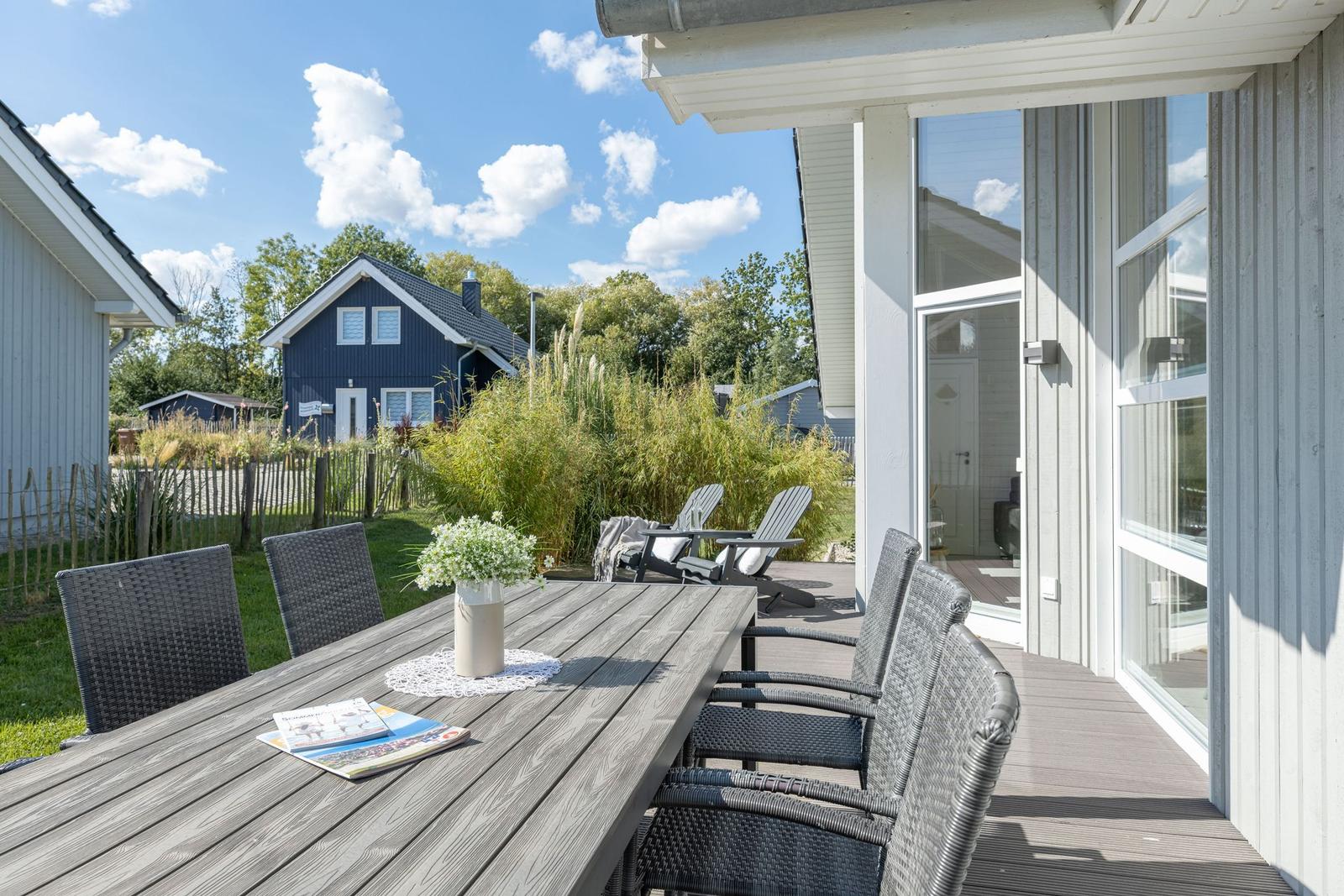 Terrasse mit Tisch und Stühlen. Im Hintergrund ein blaues Haus und Grünflächen.