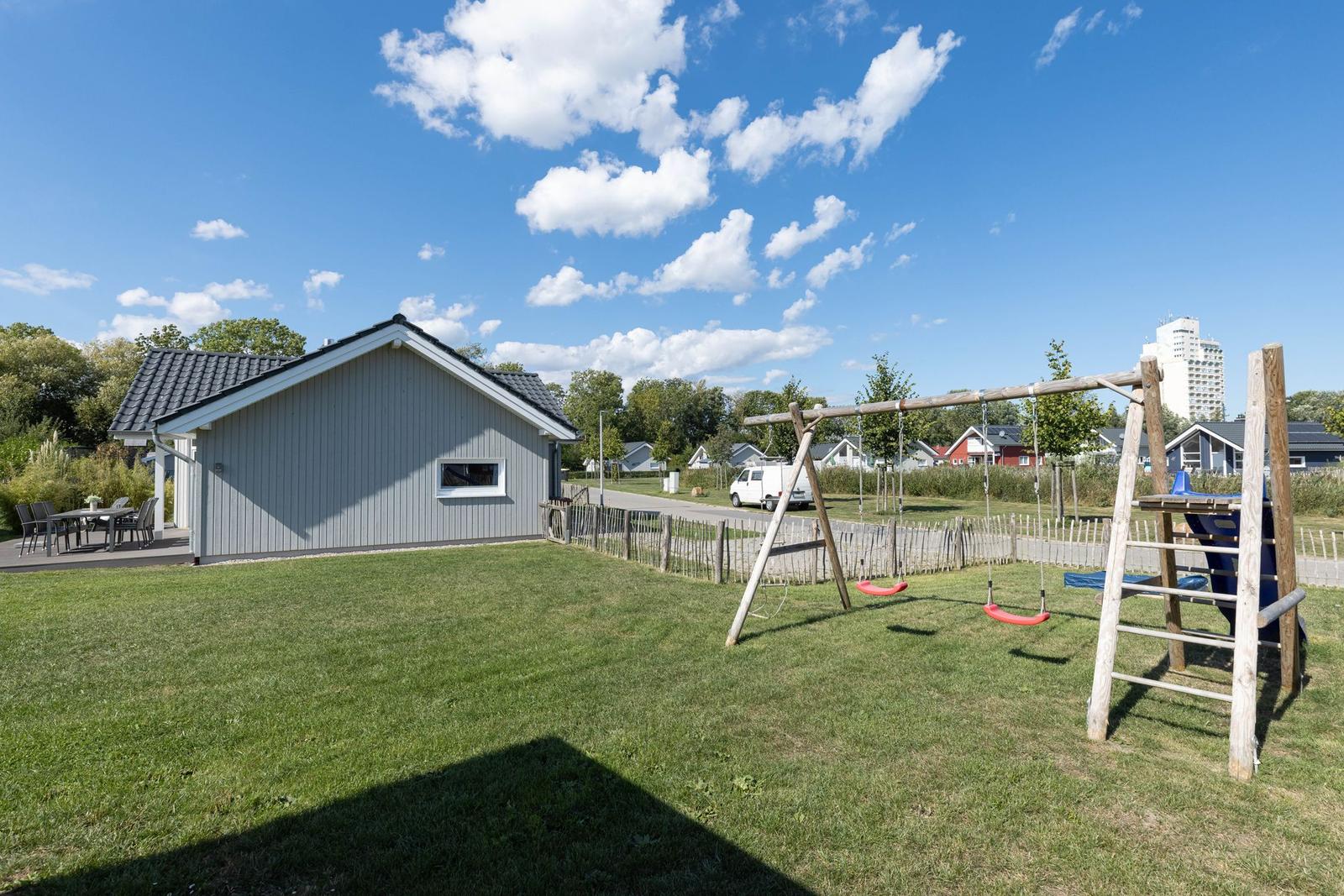 Ein graues Ferienhaus mit Garten, Spielplatz und Terrasse unter blauem Himmel.