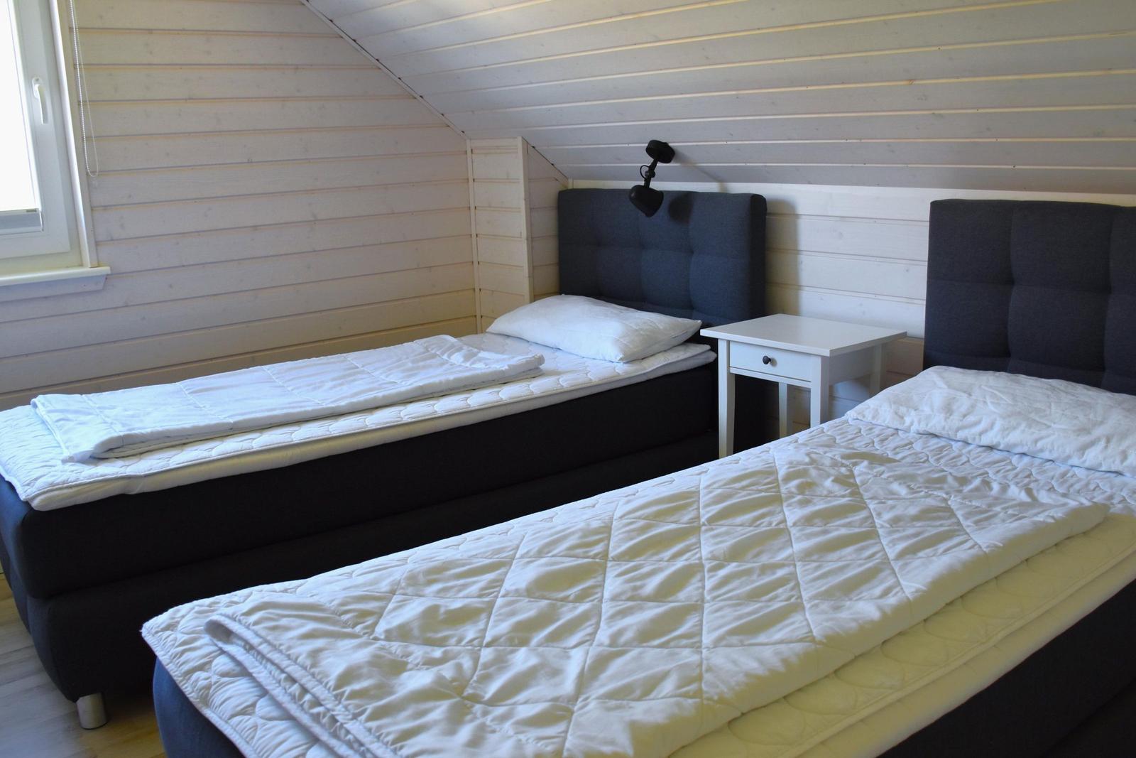 Zwei Einzelbetten mit weißen Bettwäsche und grauen Kopfenden in einem schrägen Dachzimmer mit Holzwand.