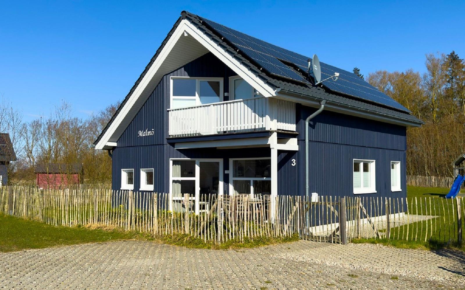Blau gestrichenes Ferienhaus mit Balkon und Solaranlage. Umzäunt mit Holzzaun und Spielplatz.