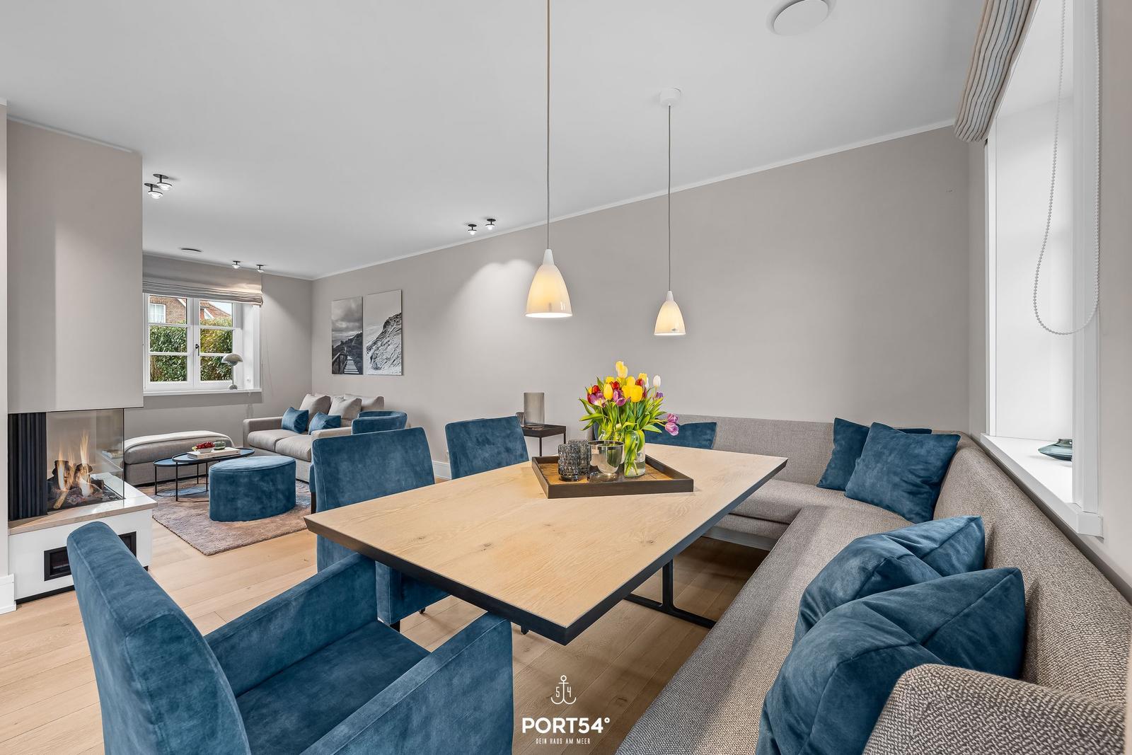 Essbereich mit Holztisch, blauen Stühlen und Sofa. Kamin und Fenster sichtbar.