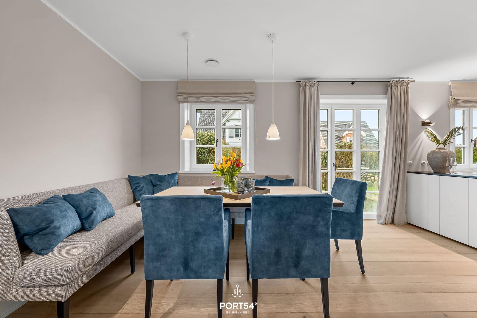 Essbereich mit Tisch, blauen Stühlen und Sofa. Fenster und Glastür zeigen Garten.