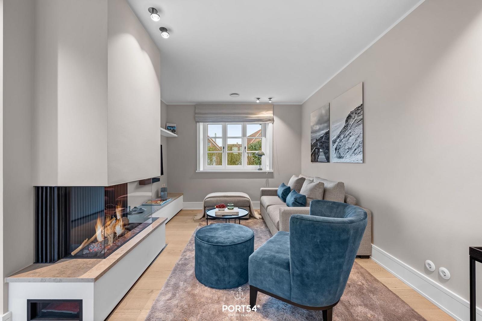 Wohnzimmer mit Kamin, Sofa und blauer Sitzgelegenheit. Fenster mit Blick nach draußen.