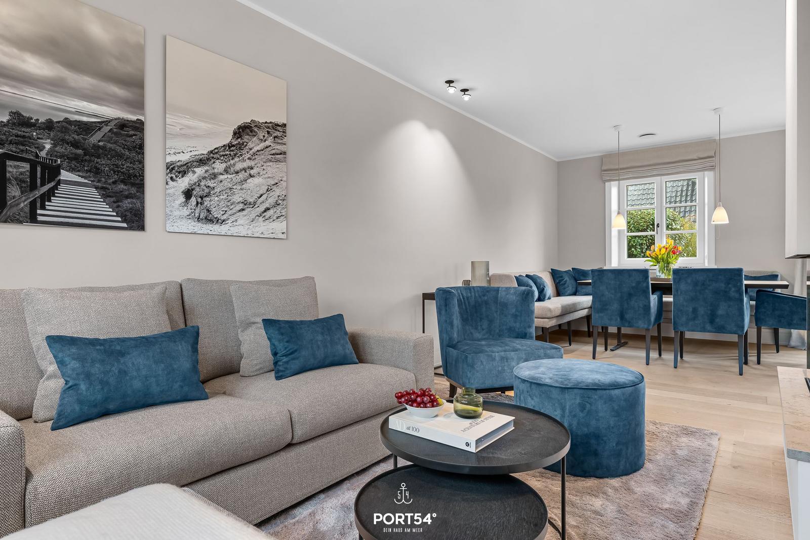 Wohn- und Essbereich mit Sofa, Sesseln und Tischen. Wandbilder und Fenster sichtbar.